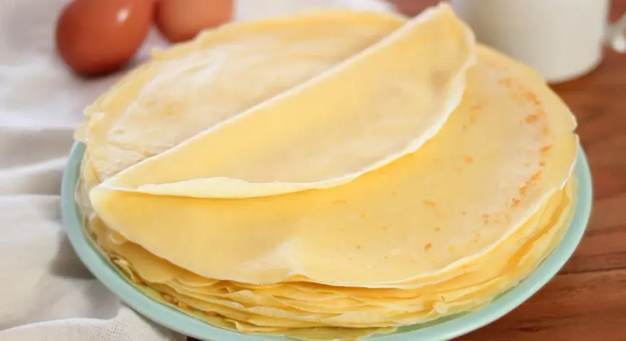Piatto di crepes senza burro, leggere e dorate, con due uova sullo sfondo.