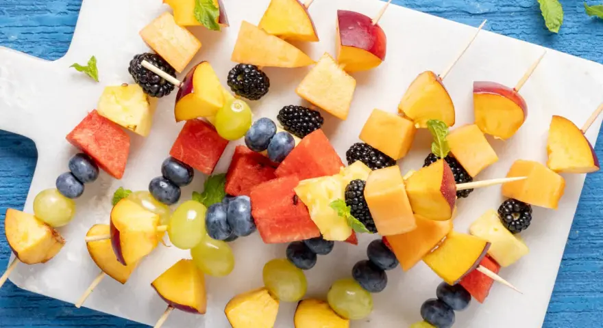 Spiedini di frutta colorati con pesche, melone, anguria, ananas, mirtilli, more e uva, guarniti con menta fresca.