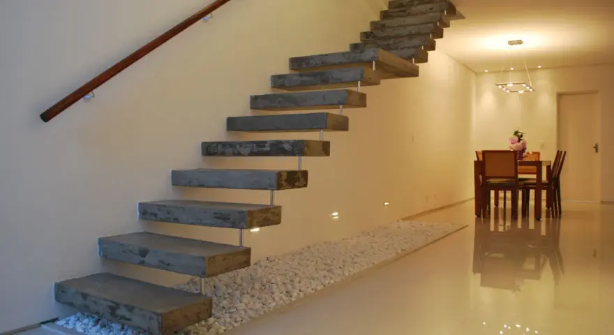 Escaleras rústicas modernas de hormigón flotante con pasamanos de madera. Un camino de guijarros blancos bordea la base.