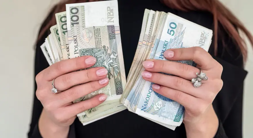 Dłonie z paznokciami w pastelowym różu trzymają pliki polskich banknotów 100 i 50 zł. To może być nagroda jubileuszowa.