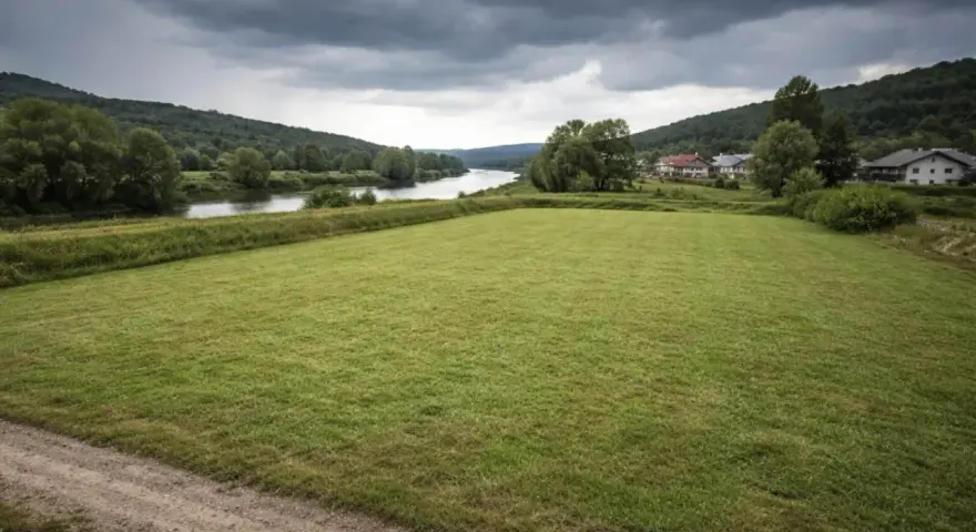 Łąka nad rzeką, domy w oddali. Chmury zapowiadają deszcz, budząc obawy o zagrożenie powodziowe.