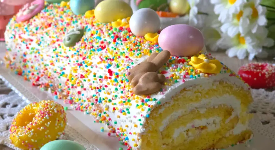 Bûche roulée décorée pour Pâques, recouverte d'un glaçage mascarpone facile et de vermicelles colorés, avec des œufs et un lapin en chocolat.