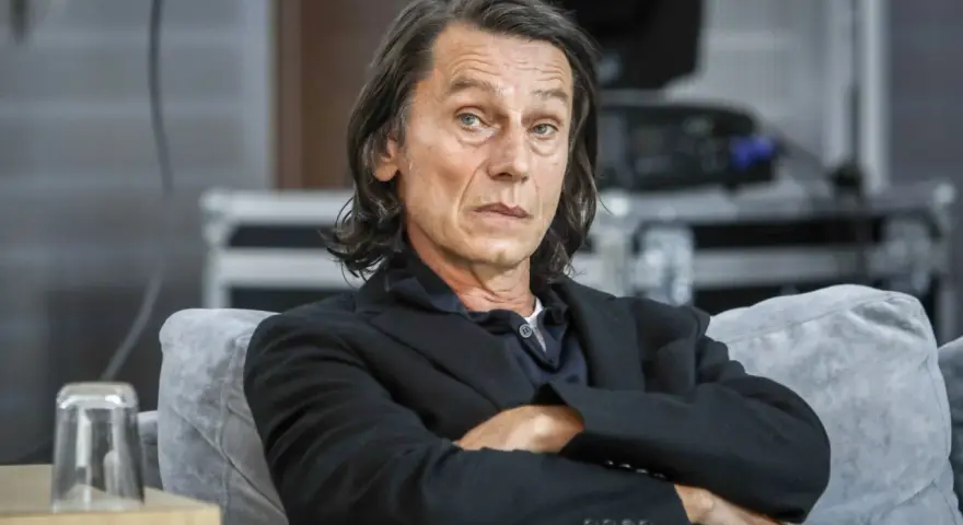 Krzysztof Warlikowski, reżyser teatralny, w czarnym garniturze, siedzi na kanapie. Jego życie prywatne budzi zainteresowanie.