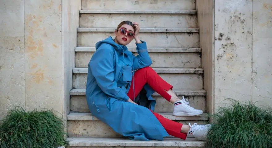Kobieta w niebieskim płaszczu i czerwonych spodniach, w białych sneakersach, prezentuje sportową elegancję na kamiennych schodach.