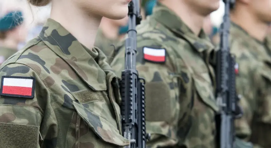 Żołnierze w mundurach z polskimi flagami, gotowi do marszu. Słychać przyśpiewki wojskowe.