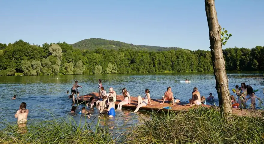 Menschen genießen einen sonnigen Tag am Baggersee Happurg. Sie sitzen auf einem Holzsteg, schwimmen und entspannen sich im Wasser.