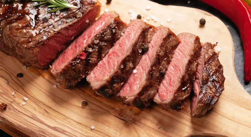 Perfekt stekt entrecote, skivad och serverad med rosmarin, pepparkorn och flingsalt på en träskärbräda. En röd chili ligger bredvid.