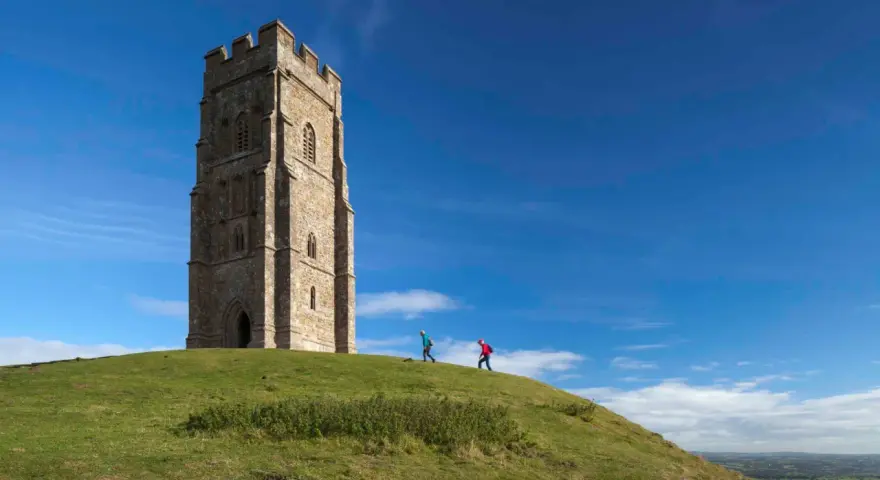 Two hikers ascend the grassy slope towards the iconic Glastonbury Tor tower under a bright blue sky.