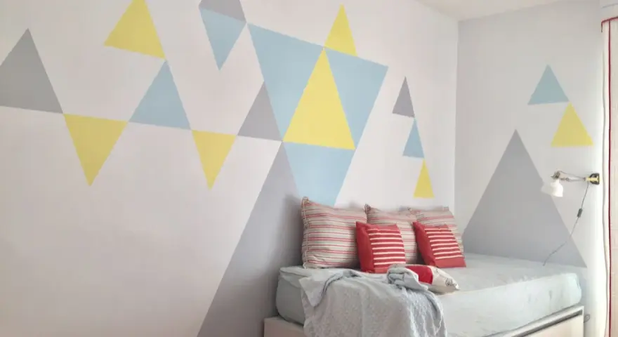 Pared pintada con triángulos de colores pasteles, creando un diseño geométrico moderno. La habitación tiene una cama con cojines y una manta.
