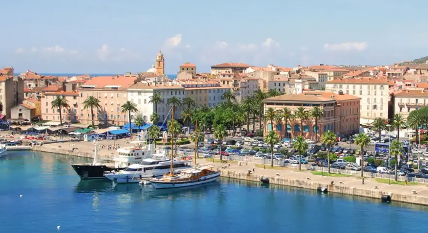 Malowniczy port na wyspie z Ajaccio, z luksusowymi jachtami i palmami, w tle kolorowe kamienice i wieża kościelna.