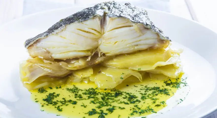 Merluza al horno con patatas, jugosa y dorada, servida sobre patatas tiernas y bañada en aceite de oliva con perejil picado.