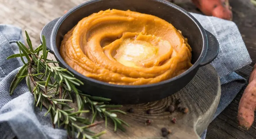 Un cremoso puré de zanahoria en un cuenco negro, coronado con mantequilla derretida. Romero fresco y granos de pimienta añaden aroma.