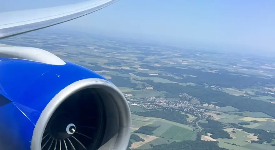 View from an airplane window, showing a blue engine and wing, with a patchwork of green fields and a small town below. Perfect for day trip flights.