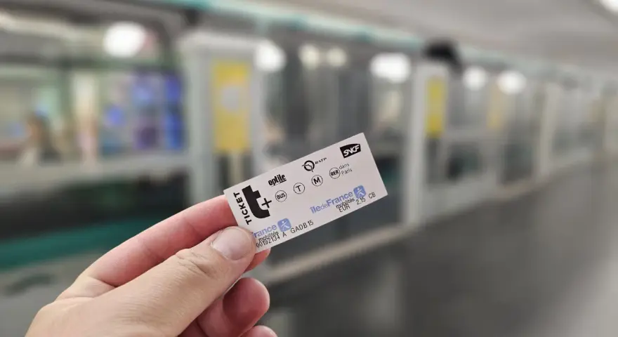 Mano sostiene un ticket t+ para el transporte público en París, con un tren borroso al fondo.