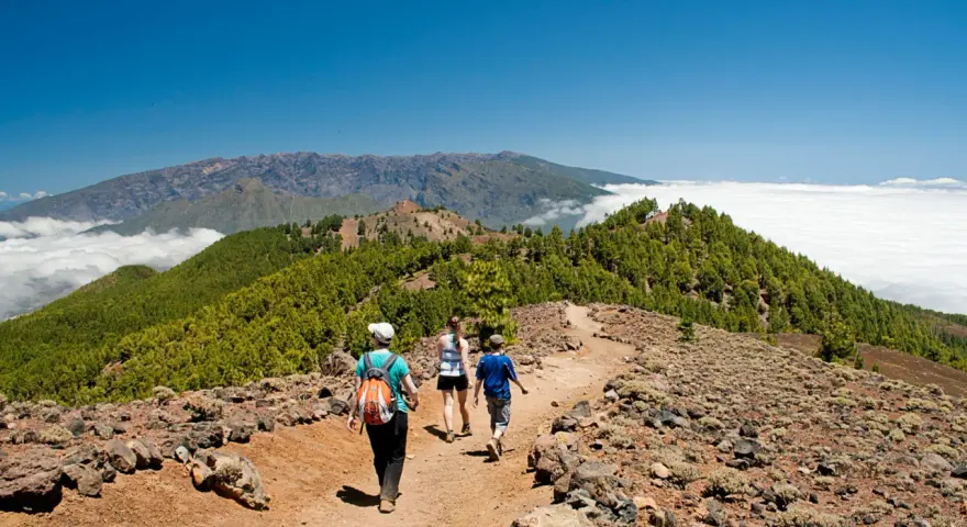 Senderos la Palma: tres personas caminan por un sendero rocoso con vistas a montañas cubiertas de pinos y un mar de nubes.
