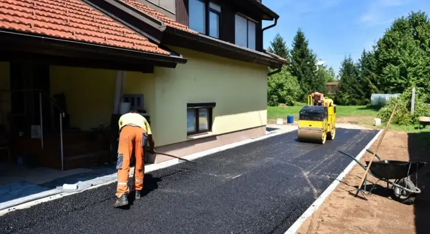 Pracownik równa łopatą świeży asfalt na podjazd, a walec go ubija. Dom w tle.