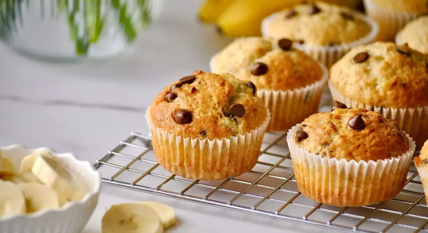 Frisch gebackene bananen muffins mit Schokostückchen auf einem Gitter, daneben eine Schüssel mit Bananenscheiben.