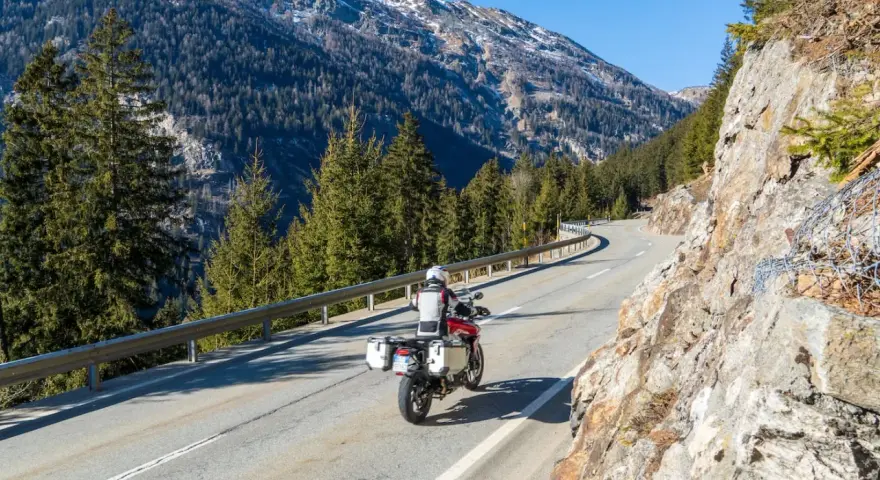 Motociclista su strada alpina in Svizzera, con montagne innevate e foreste. Ottimi consigli per un viaggio in moto in Svizzera.