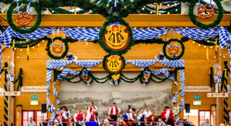 Wnętrze namiotu na Oktoberfest ozdobione niebiesko-białymi girlandami i wiankami z precelkami. Ludzie siedzą przy stołach, a w tle widać orkiestrę.