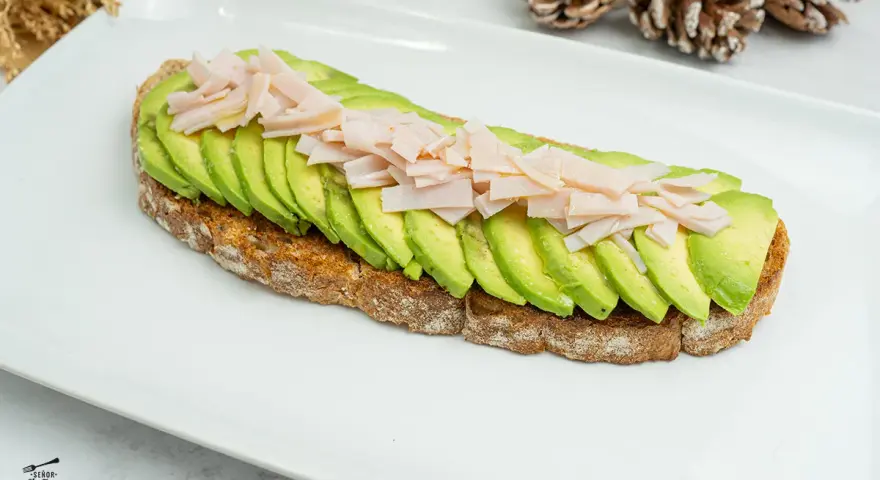 Tostada integral con aguacate en rodajas y jamón picado sobre plato blanco.