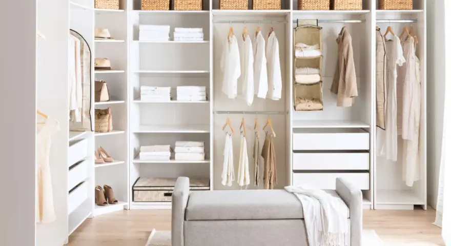 A bright, organized closet with white DIY closet shelves, featuring neatly folded clothes, baskets, and hanging garments. A grey bench sits in front.