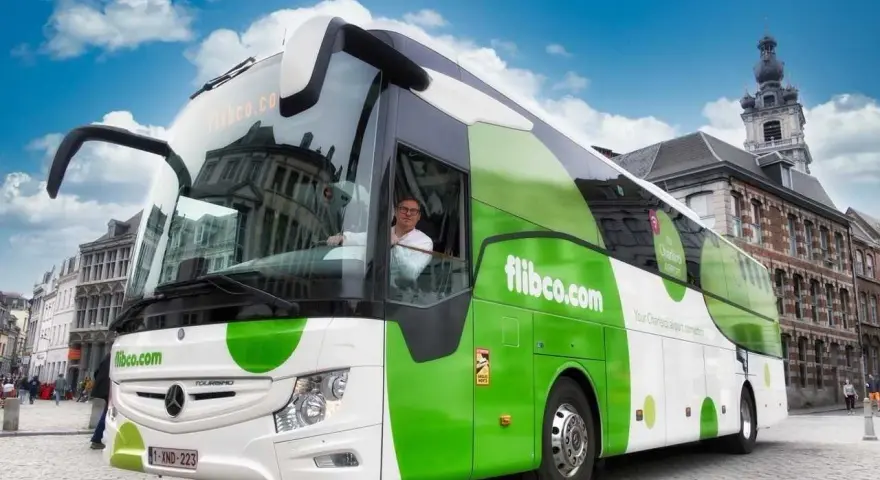 Autobus flibco.com z kierowcą, na tle miejskiej zabudowy i błękitnego nieba.