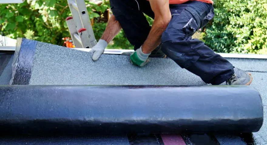 Handwerker verlegt Dachschindeln. Eine Rolle Bitumenbahn liegt bereit, während die neue Schicht auf dem Dach ausgebreitet wird.