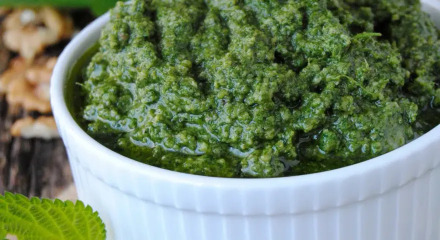 Pesto z pokrzywy w białej miseczce, obok orzechów włoskich i świeżych liści pokrzywy.