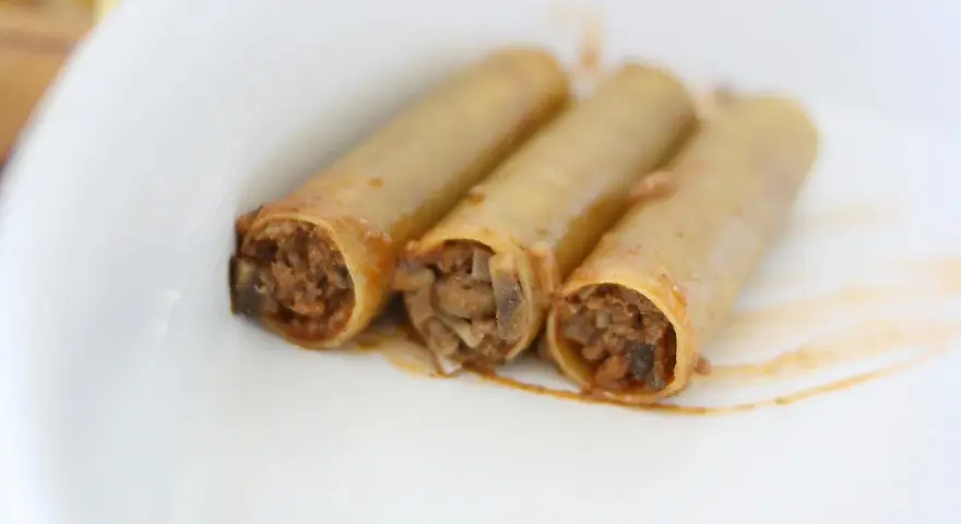 Trzy cannoloni z farszem mięsnym, wyglądające jak naleśniki z mięsem, leżą na białym talerzu.