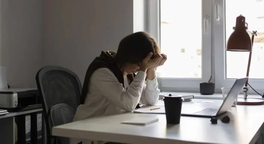 Une personne accablée par la pression au travail, la tête entre les mains, assise à un bureau encombré.