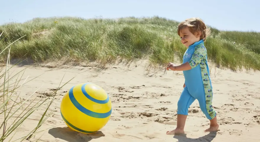 Chłopiec w niebieskim stroju kąpielowym z neoprenu bawi się żółtą piłką na plaży.