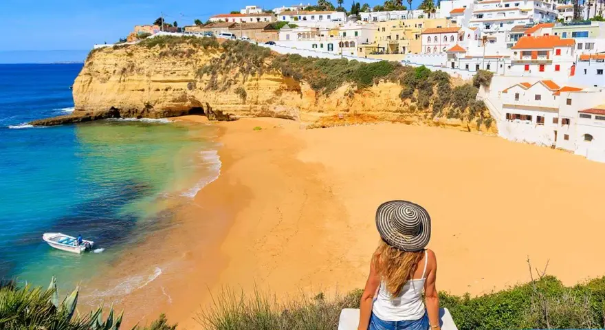 A woman in a sun hat enjoys the view of a golden beach and turquoise water in Portugal's Algarve region.
