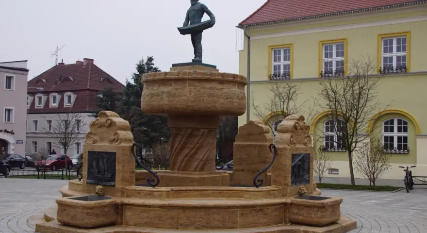 Kamienna fontanna z rzeźbą rybaka na szczycie, ozdobiona płaskorzeźbami statków.