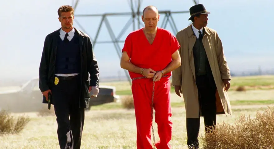 Tre uomini camminano in un campo: un detenuto in tuta rossa ammanettato, un poliziotto e un detective. Scena da film polizieschi recenti.
