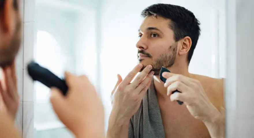 Hombre con barba corta se afeita la barba con una recortadora, buscando un afeitado apurado frente al espejo.