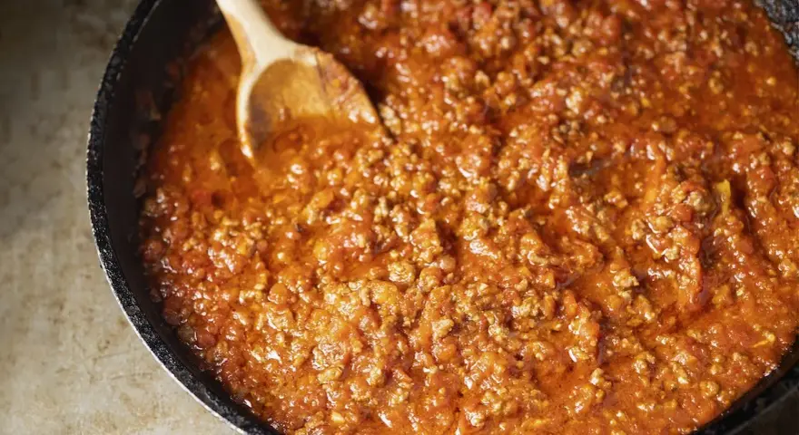 Un saporito ragù di carne in una padella, pronto per essere servito. Un cucchiaio di legno mescola la salsa ricca e densa.