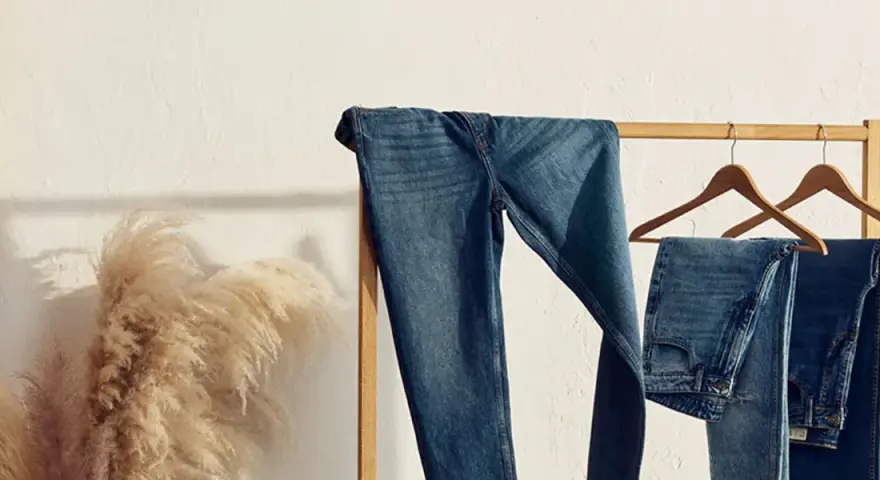 Varios pares de pantalones de **tejido denim** cuelgan de una percha de madera. A la izquierda, un gran ramo de hierba de la pampa añade un toque natural.