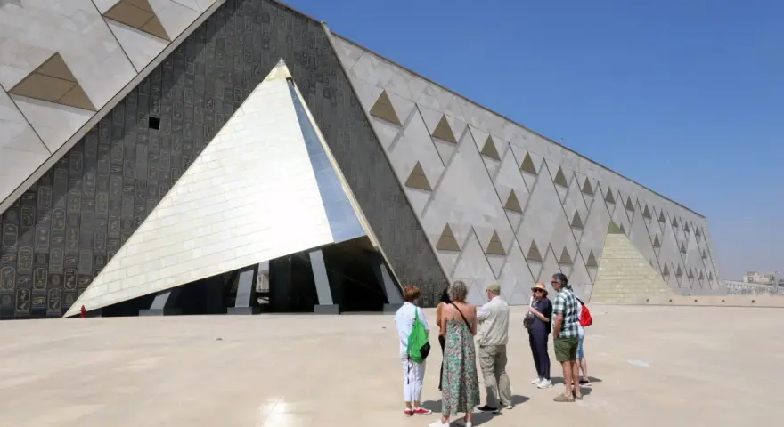 Grupa turystów podziwia nowoczesne muzeum w Gizie, którego fasada zdobiona jest hieroglifami i geometrycznymi wzorami przypominającymi piramidy.