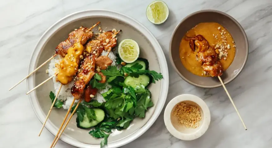 Pyszny kurczak satay na ryżu z orzeszkami ziemnymi, ogórkiem i limonką. Obok miseczka z sosem orzechowym.