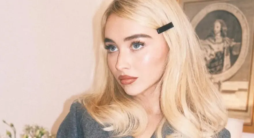 Blondynka z **side part fryzurą** i czarną spinką, w szarym żakiecie, z błękitnymi oczami.