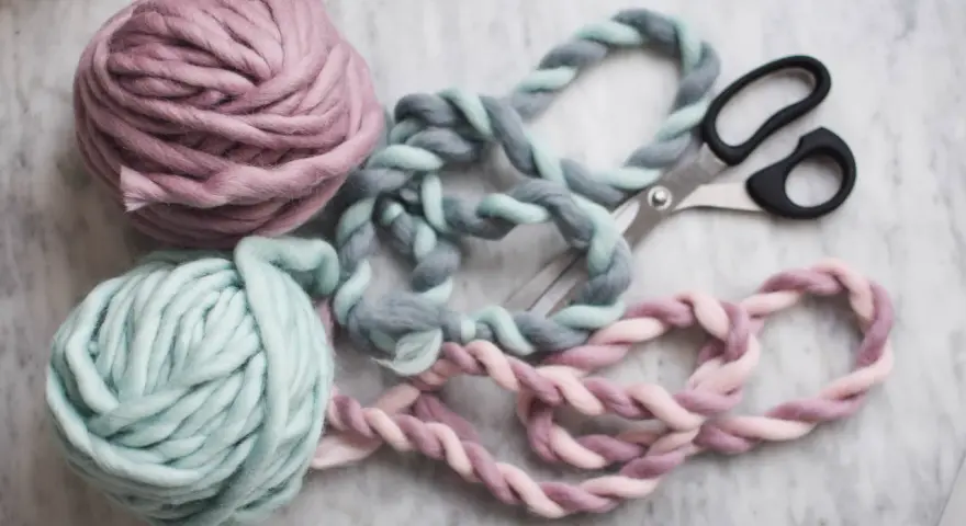 Två härvor tjockt garn i pastellfärger, en tvinnad tråd i grått och mintgrönt, samt en i rosa och ljusrosa, ligger bredvid en sax på en marmorerad yta.