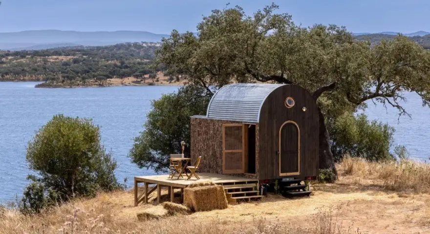 Une caravane transformée en cabane insolite, avec une terrasse en bois, au bord d'un lac paisible.