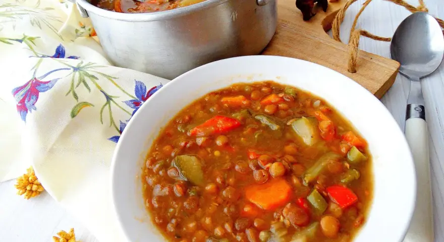 Guiso de lentejas con verduras en un cuenco blanco, junto a una olla y servilleta floral.