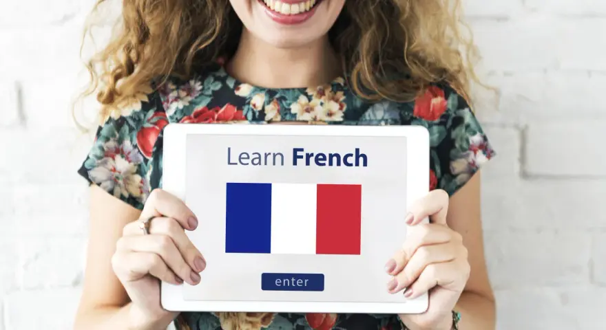 Una mujer sonriente sostiene una tablet que muestra "Learn French" y la bandera francesa. ¡Es hora de aprender como se habla en Francia!