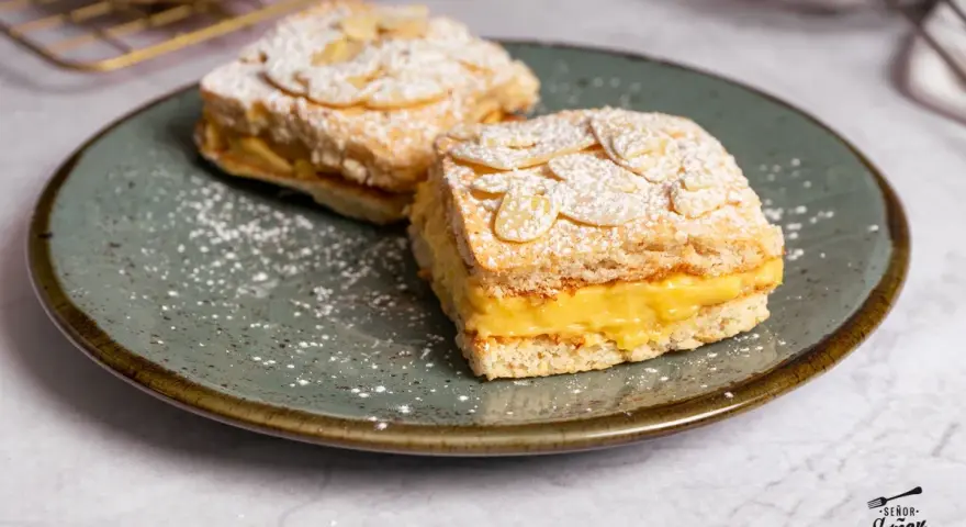 Dos porciones de pastel ruso con crema amarilla y almendras laminadas, espolvoreados con azúcar glas.
