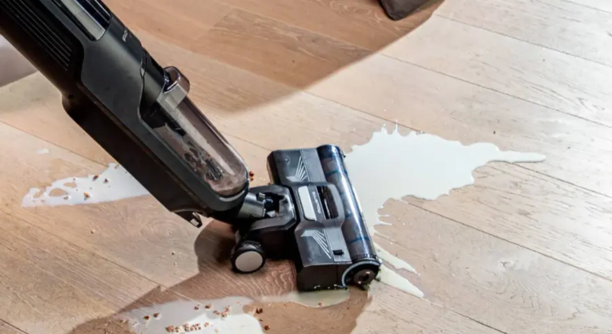 A wet/dry vacuum cleaner is cleaning up a spill of white liquid and debris on a wooden floor.