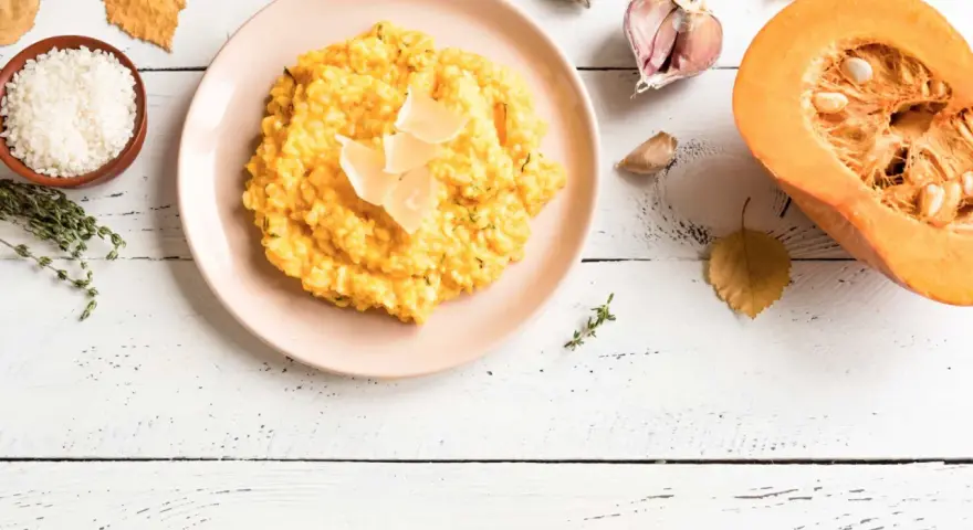 Un cremoso risotto de calabaza, decorado con láminas de queso, junto a ingredientes frescos como arroz, tomillo, ajo y una calabaza partida.
