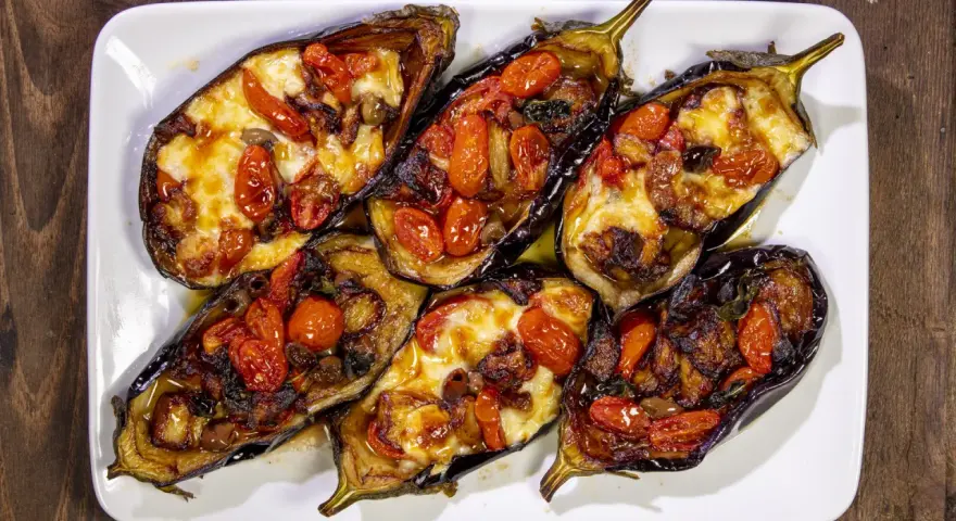 Melanzane a scarpone ripiene di pomodorini, formaggio filante e olive, cotte al forno su un piatto bianco.