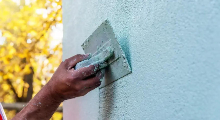 Ręka z kielnią wykonuje tynkowanie ścian. Jasne, jesienne liście w tle tworzą ciepłą atmosferę.