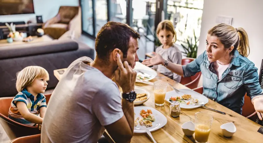 Familie am Esstisch, die Frau erklärt gestikulierend, wie streitet man richtig, während der Mann sich die Ohren zuhält.
